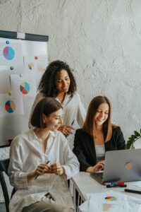 Tres mujeres emprendedoras sonríen frente a un ordenador mientras analizan gráficas de marketing digital en una pizarra, representando trabajo en equipo y crecimiento online.